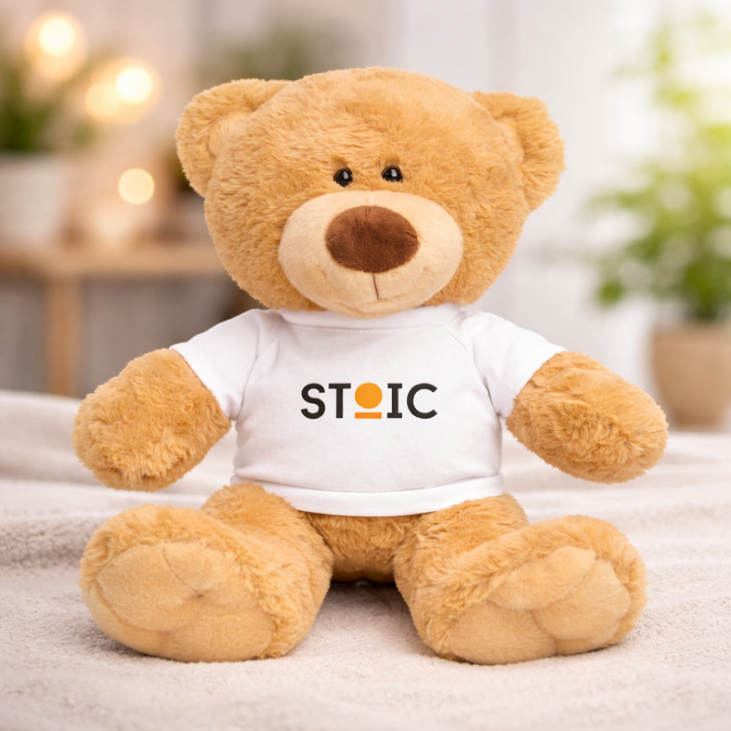 Teddy bear wearing a white shirt with 'STOIC' logo on a blurred indoor background