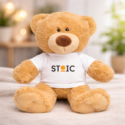 Teddy bear wearing a white shirt with 'STOIC' logo on a blurred indoor background