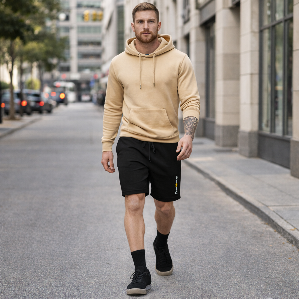 Man walking on a city street wearing a beige hoodie and black shorts with embroidered STOIC brand logo vertical, and black sneakers.